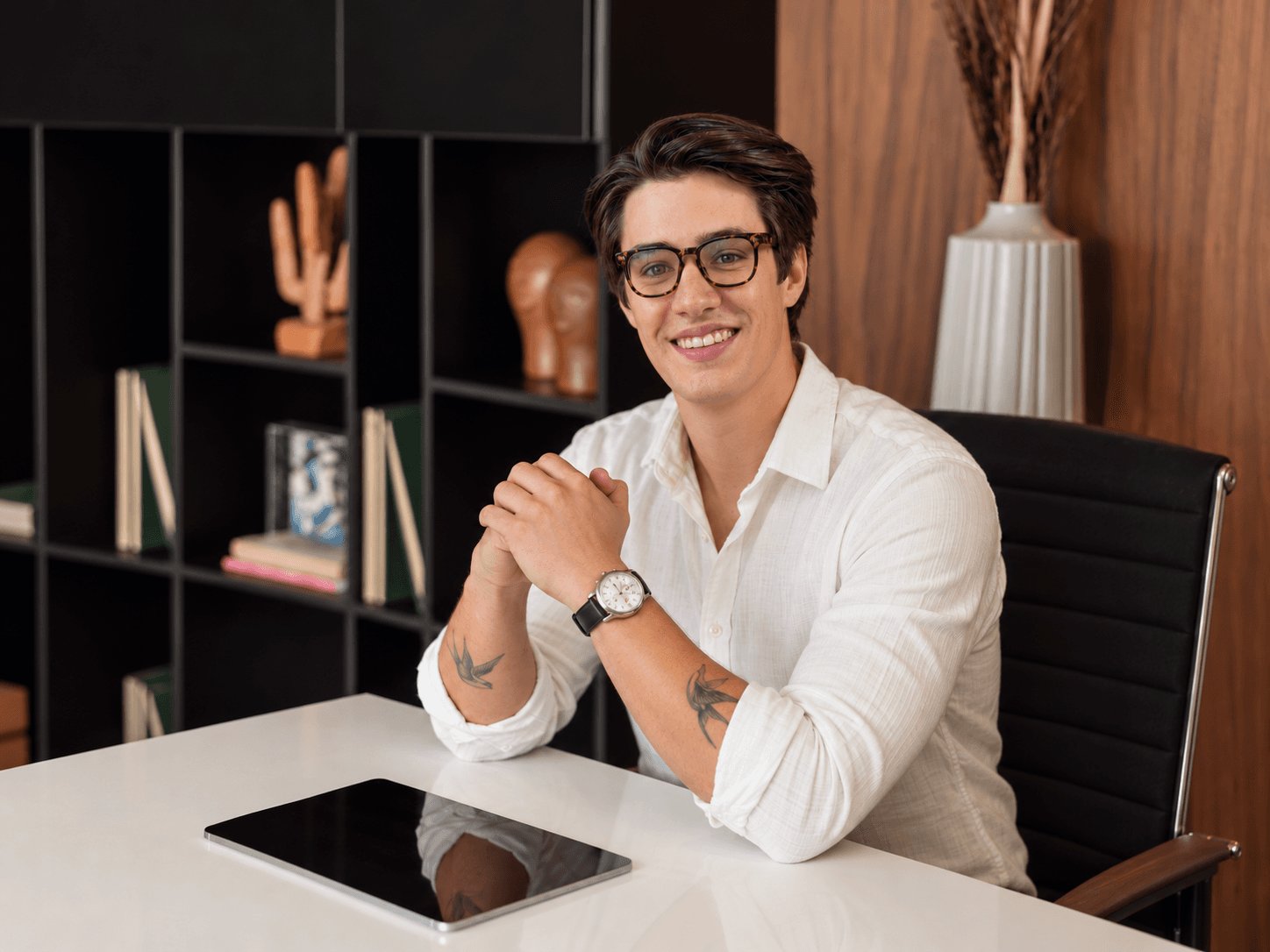 Cauê Resina, sócio da Studio Side, sorrindo sentado à mesa do escritório, diante de uma estante preta e de um painel de madeira