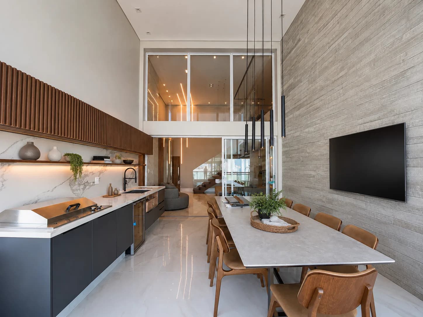 Cozinha com bancada escura e mesa de jantar longa sob o pé-direito duplo, com vidro do piso ao teto ao fundo