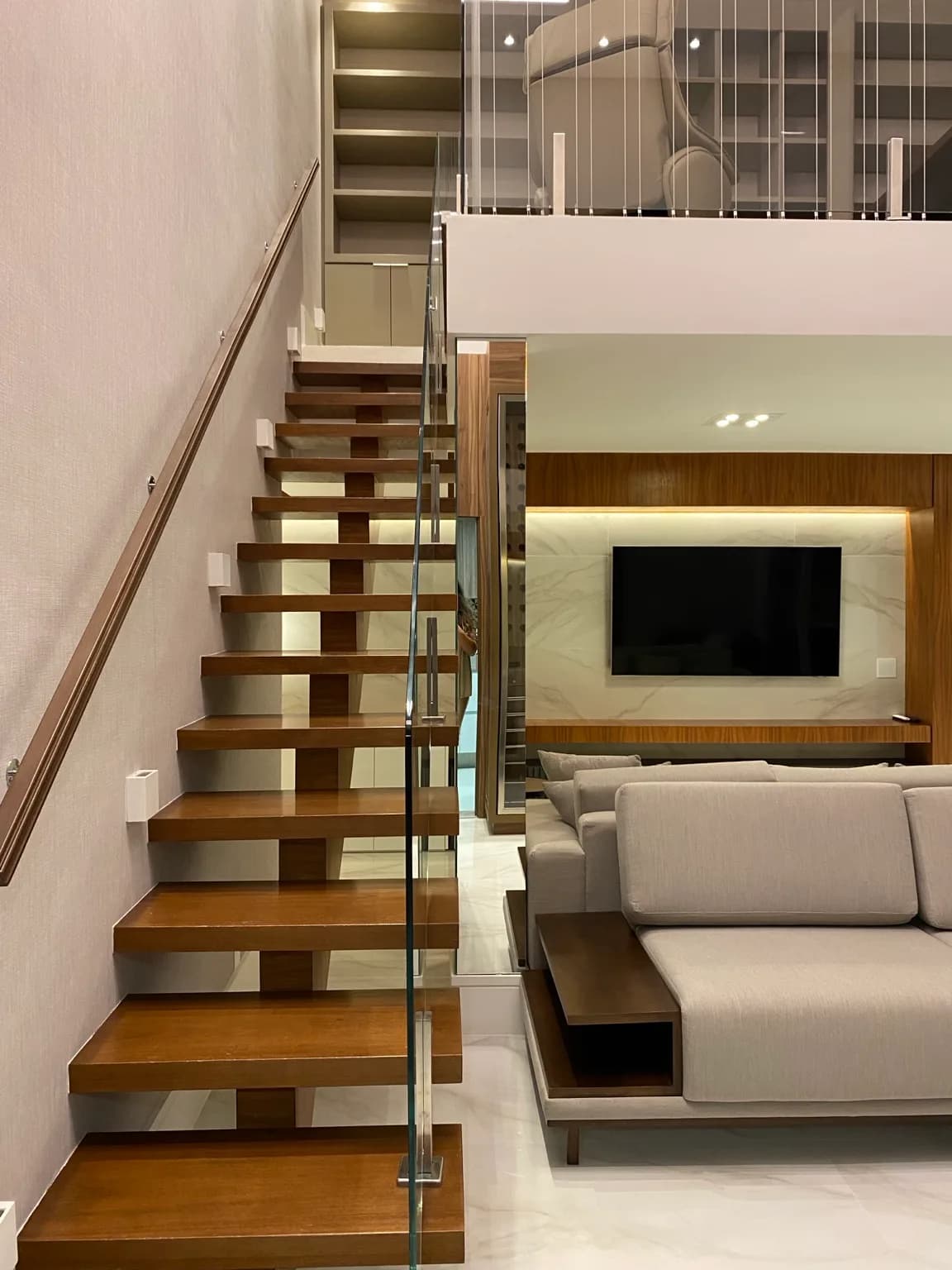 Escada de degraus de madeira com guarda-corpo de vidro, com a sala e o painel de TV ao fundo