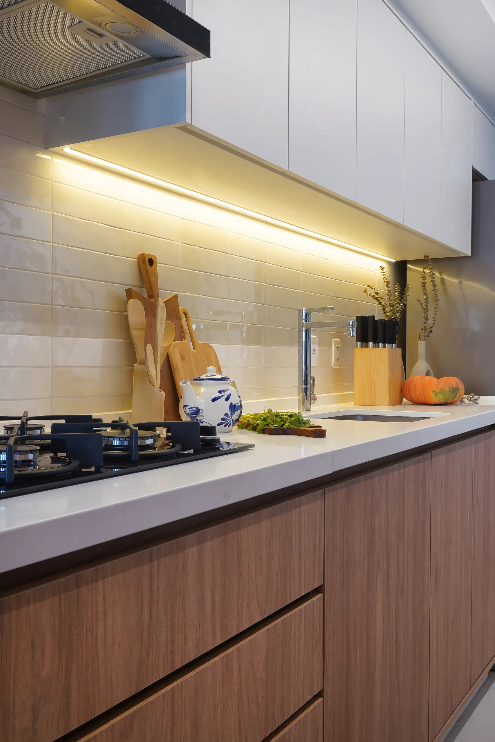 Bancada da cozinha em uso, com cooktop, tábuas de madeira, temperos e iluminação sob os armários