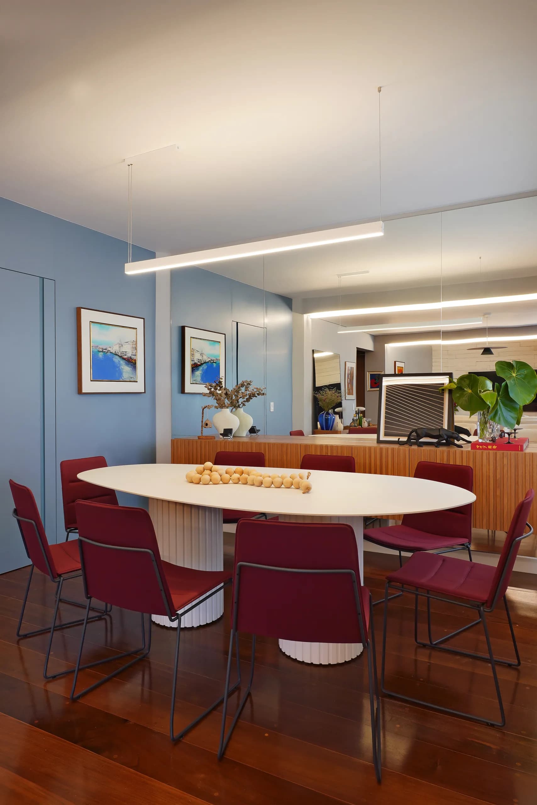 Mesa de jantar oval branca com cadeiras bordô, sobre piso de madeira e parede azul