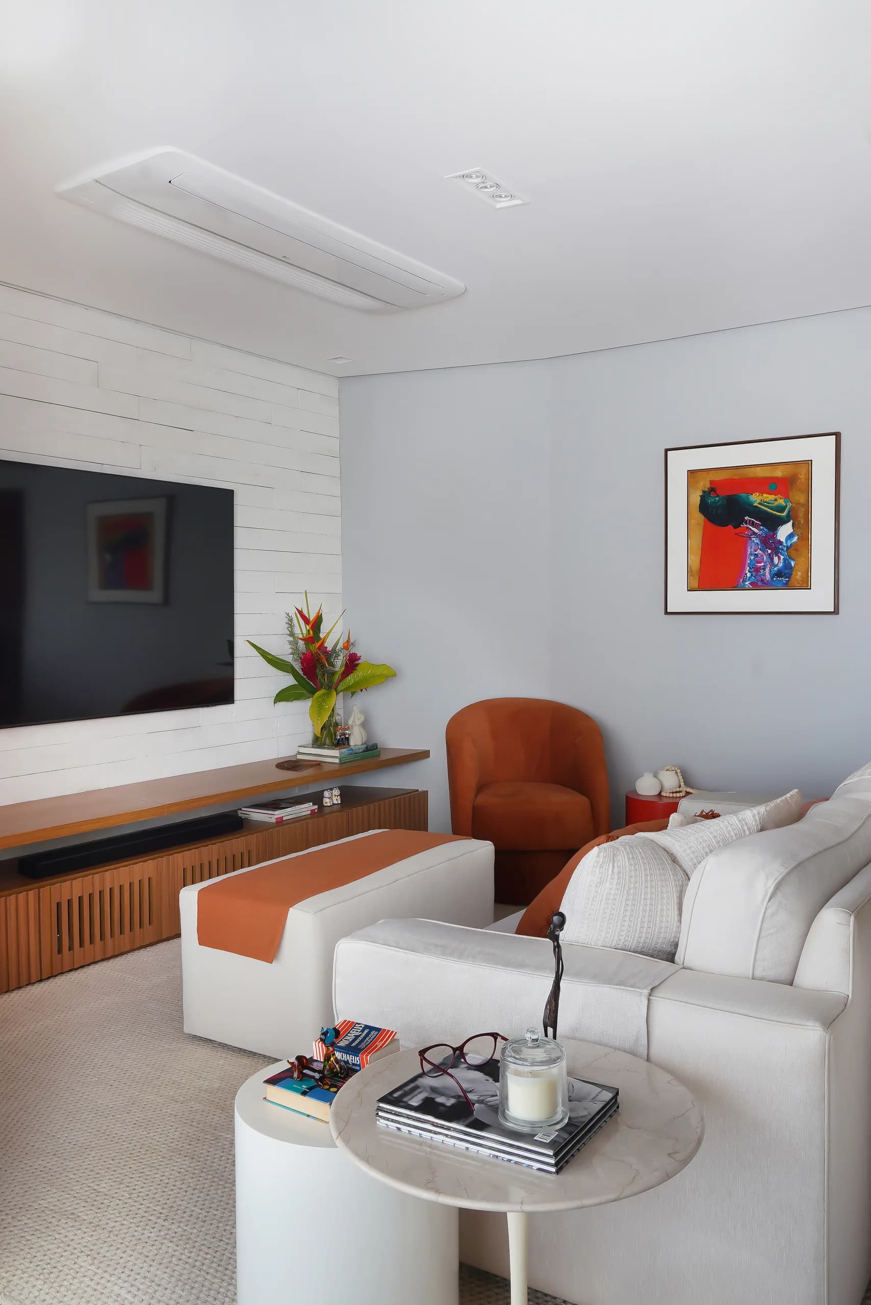 Sala com TV na parede de tijolo branco, rack de madeira ripada, sofá claro, pufe com manta laranja e poltrona terracota
