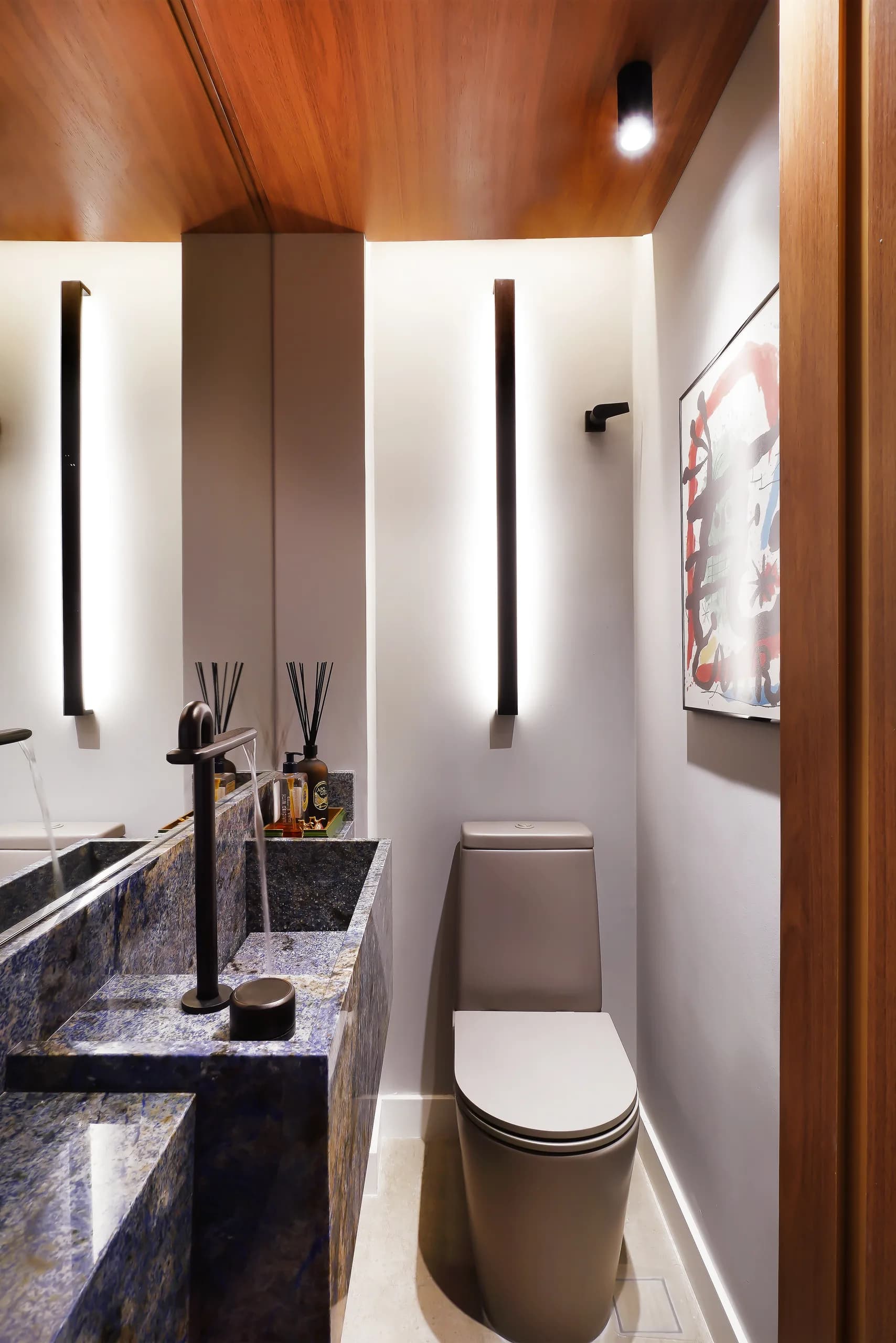 Lavabo com bancada de pedra escura, arandelas verticais e marcenaria de madeira